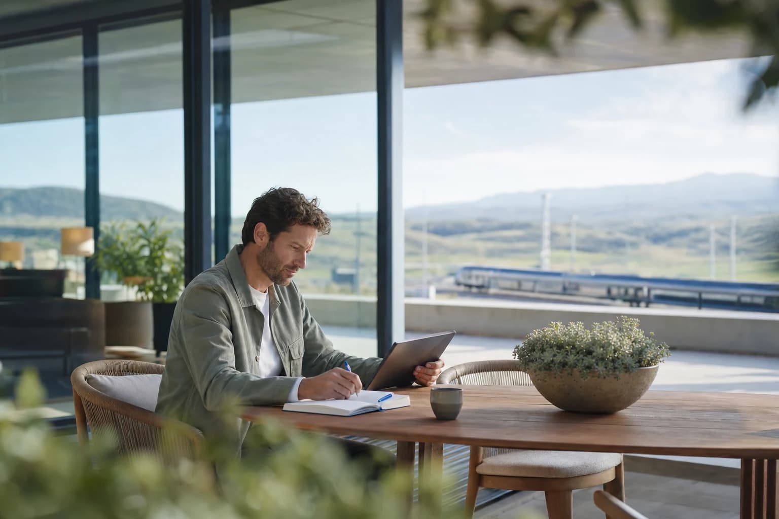 A researcher works with a tablet and notebook in a bright lounge overlooking an open landscape.